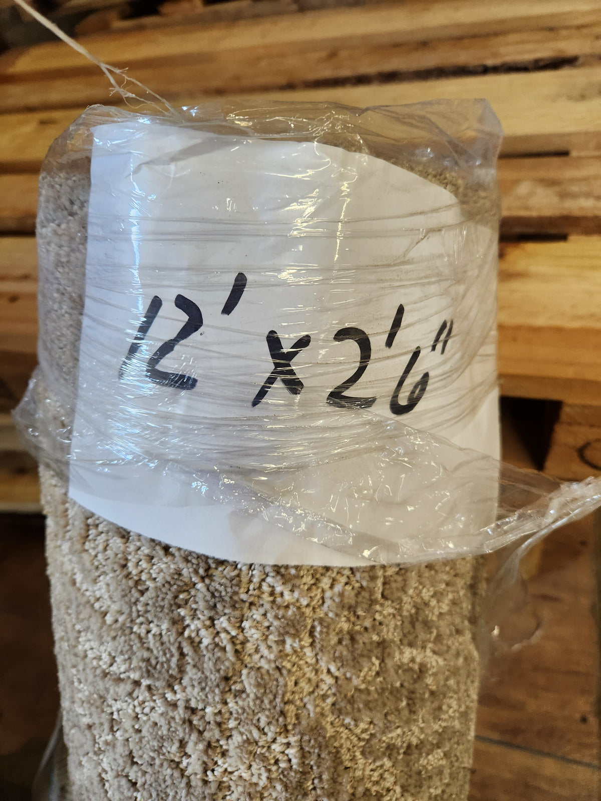 A Flooring CLEARANCE Carpet Remnant 12' X 2'6" in beige/grey, rolled in plastic; wood planks seen behind.