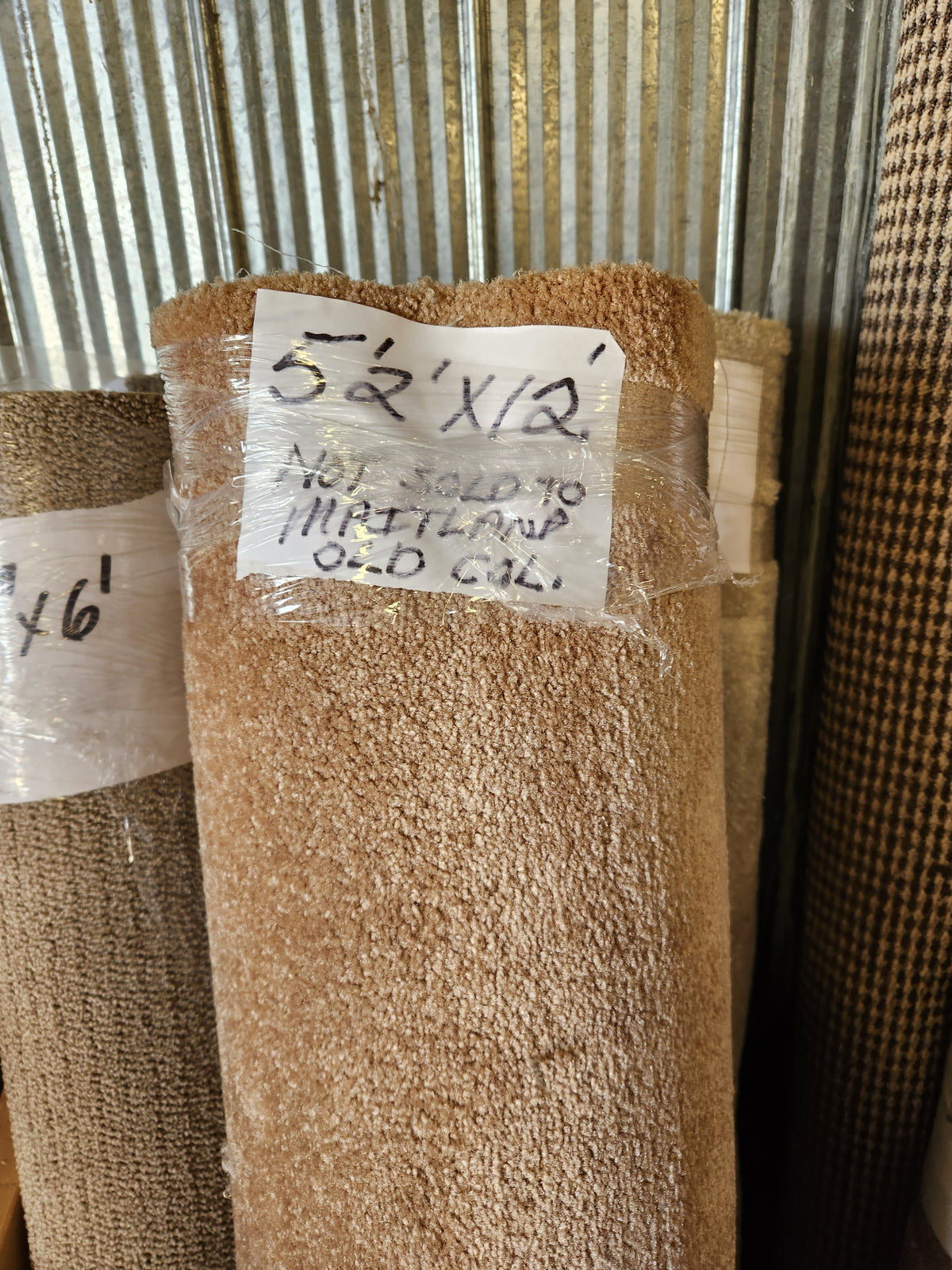A Flooring CLEARANCE Carpet Remnant 5'2" x 12', beige and wrapped in plastic, stands by a metal wall with notes.