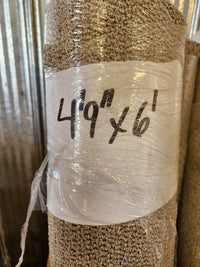 A Flooring CLEARANCE Carpet Remnant 4'9" x 6', wrapped in plastic, leans against a corrugated metal wall.