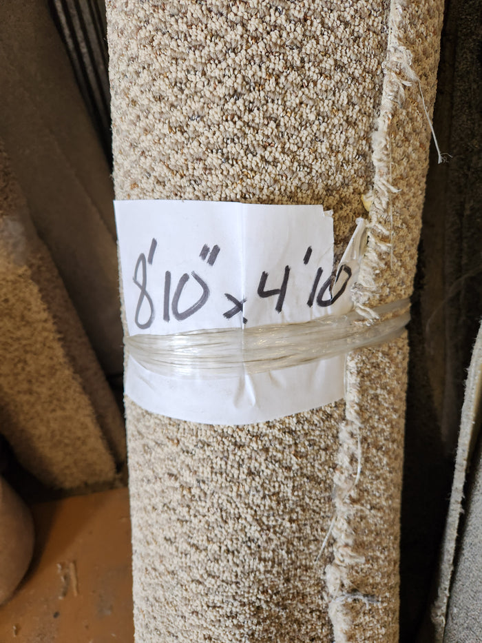 A Flooring CLEARANCE Carpet Remnant 8'10" x 4'10", tan low pile, rolled with a handwritten sign taped around it.