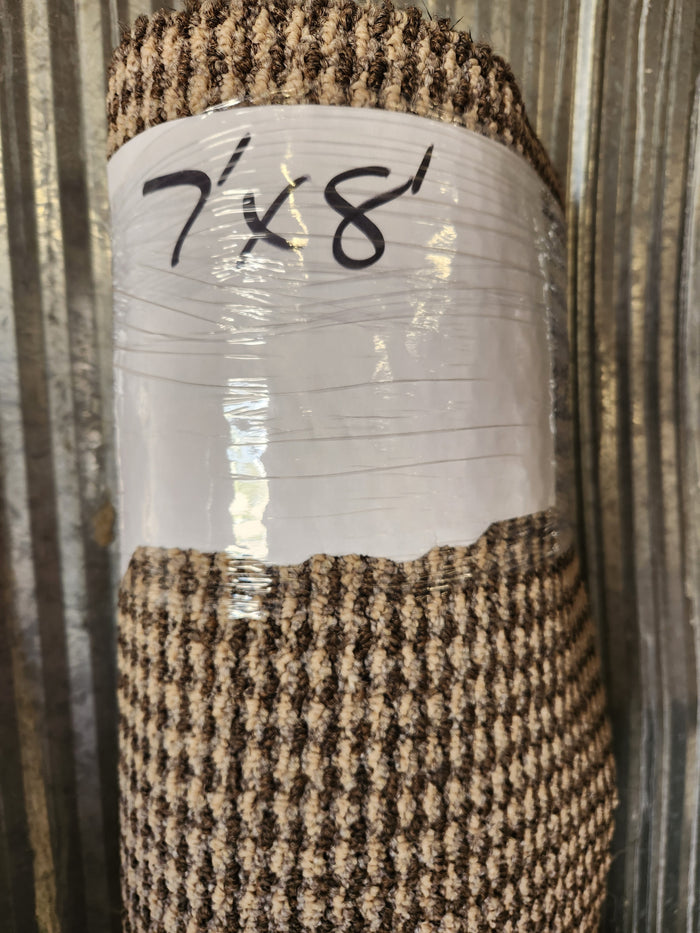 A Flooring CLEARANCE Carpet Remnant 7' X 8', brown/beige, low pile, wrapped in plastic against a metal wall.