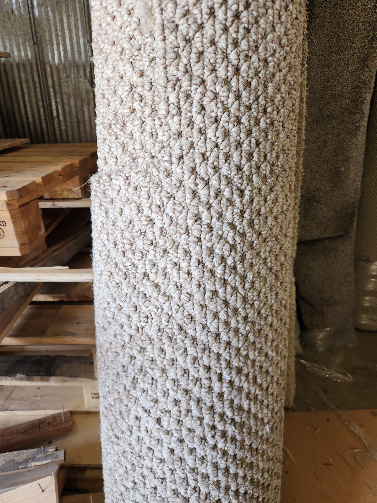 A Flooring CLEARANCE Carpet Remnant 12' X 3'9" in beige stands upright by wooden pallets in an industrial space.
