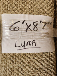 A beige low pile Carpet Remnant 6' x 8'7" by Flooring CLEARANCE, labeled with marker on paper and plastic wrap.