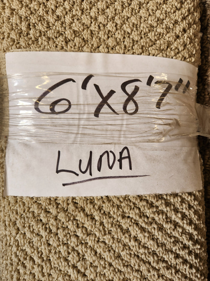 A beige low pile Carpet Remnant 6' x 8'7" by Flooring CLEARANCE, labeled with marker on paper and plastic wrap.