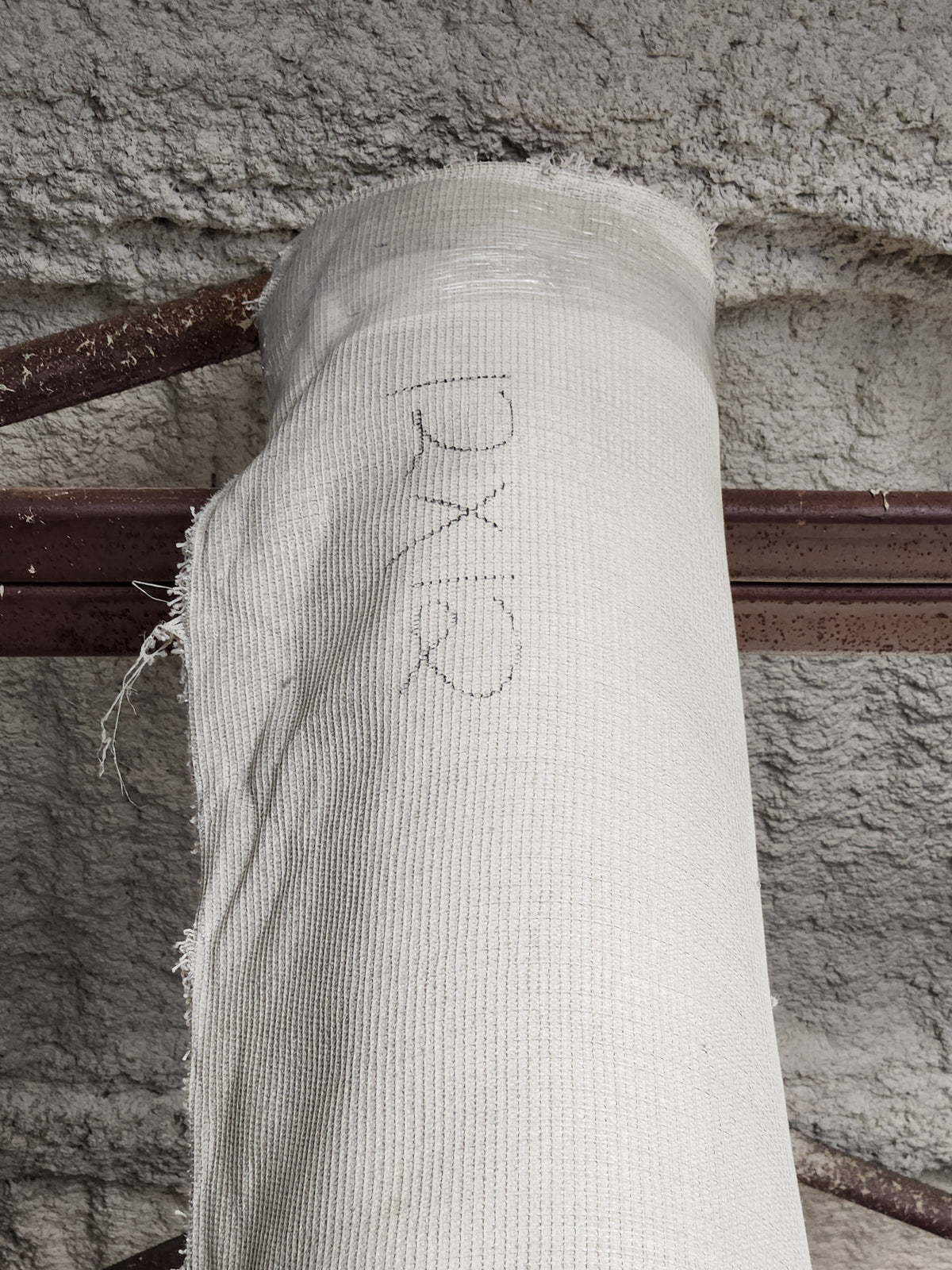 A Flooring CLEARANCE Carpet Remnant 12' X 12' in off-white rests on a rusted beam against a textured wall.