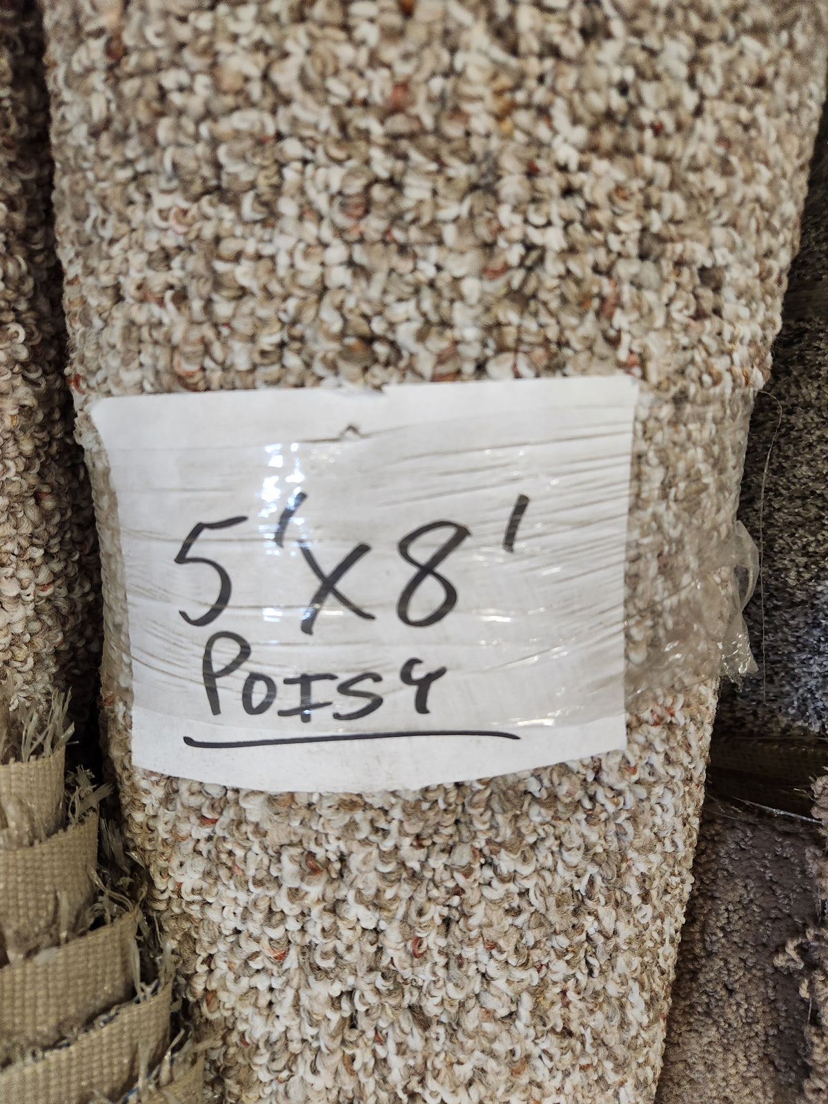 A rolled Carpet Remnant 5' X 8' by Flooring CLEARANCE with a handwritten sign taped to the beige carpet.