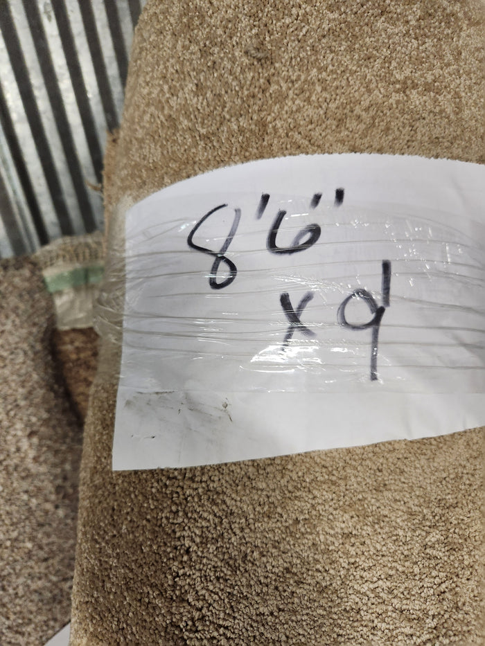 A roll of Flooring CLEARANCE Carpet Remnant 8'6" x 9', beige, medium pile, with a white handwritten label.