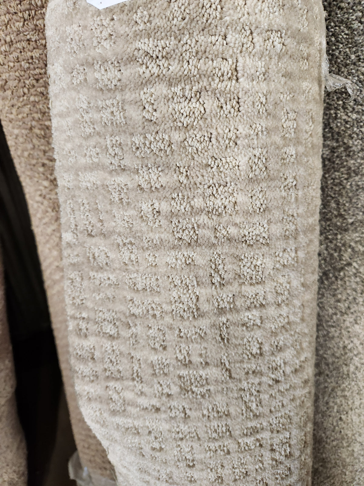 A close-up of Flooring CLEARANCE Carpet Remnant 8'6" X 9'4", tan grid-pattern, stacked with other rolls.