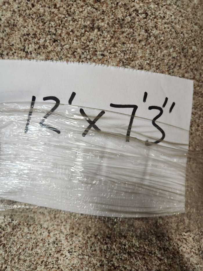 A Carpet Remnant 12' X 7'3" by Flooring CLEARANCE, light brown, lies wrapped in plastic with a handwritten note.