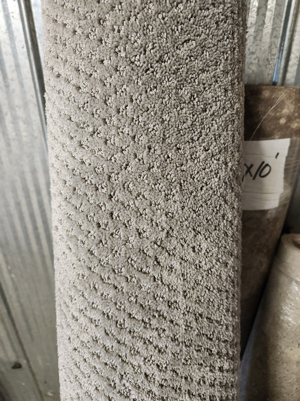 A Flooring CLEARANCE Carpet Remnant 6' X 10' in grey stands upright by another roll near a corrugated metal wall.
