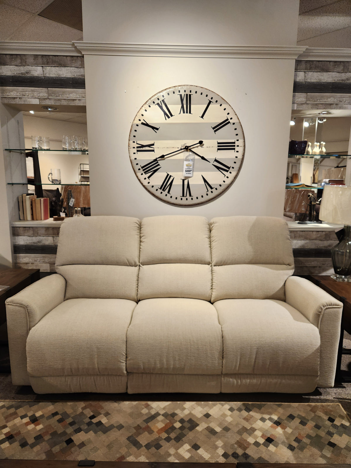 A La-Z-Boy 803 Cade Manual Reclining Sofa in beige sits below a Roman numeral clock in a showroom.