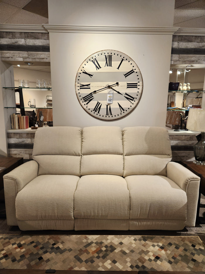 A La-Z-Boy 803 Cade Manual Reclining Sofa in beige sits below a Roman numeral clock in a showroom.
