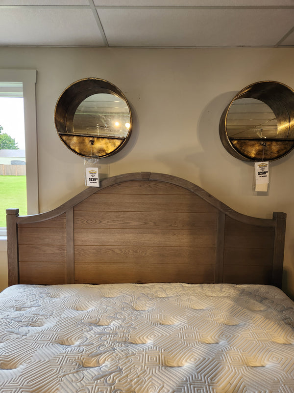 The Vaughan Bassett 740 Bungalow Arch King Bed with a patterned comforter sits beneath two round mirrors in a bright room.