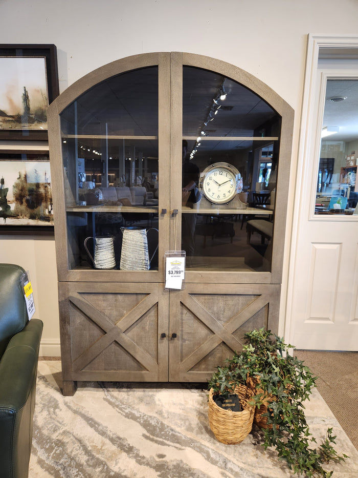The Barrett Cabinet by Mercana features glass doors, displaying a clock and metal pitchers; potted plant in front.