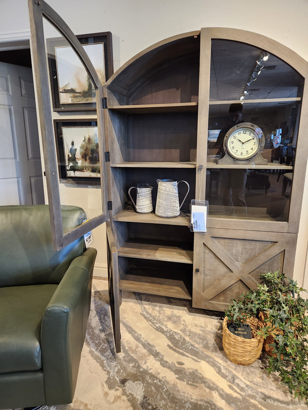 Mercana Barrett Cabinet with glass doors, shelves for pitchers and clock, next to green armchair and potted plant.