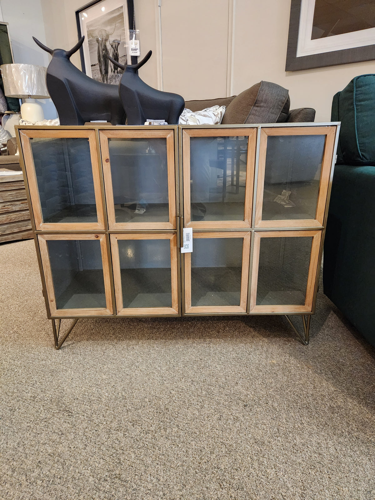 The Pandora Accent Cabinet by Mercana features metal legs and glass doors, shown in a furniture store showroom.
