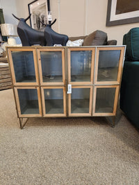 The Pandora Accent Cabinet by Mercana features metal legs and glass doors, shown in a furniture store showroom.