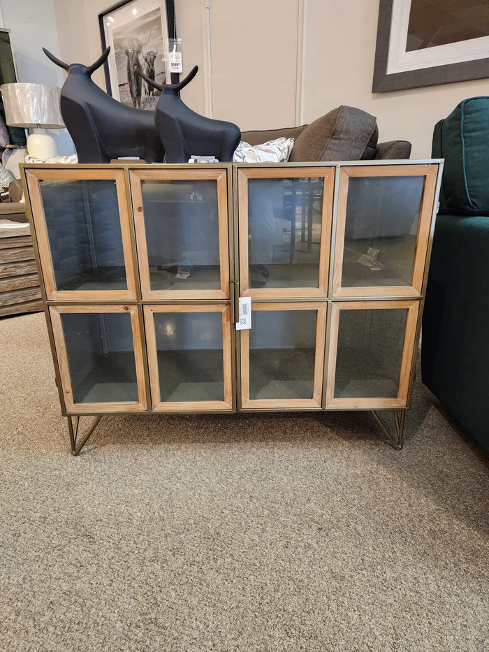 The Pandora Accent Cabinet by Mercana features metal legs and glass doors, shown in a furniture store showroom.