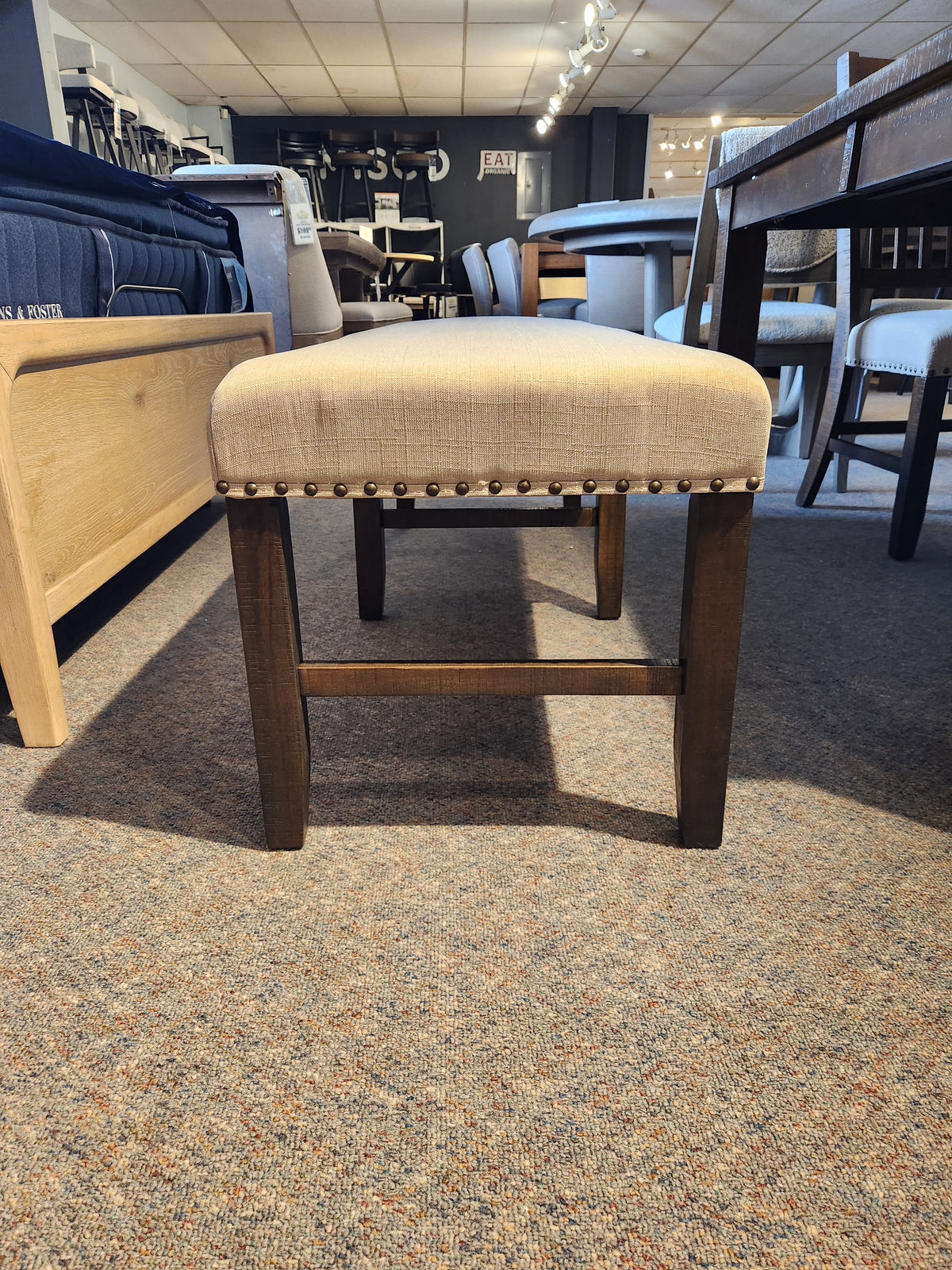 The Willow Creek Bench by Jofran features a backless beige seat, nailhead trim, and dark wood legs for rustic style.