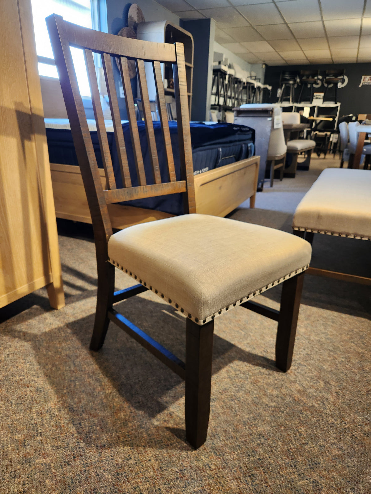 The Willow Creek Slatback Chair by Jofran features a beige upholstered seat, displayed in a modern furniture showroom.