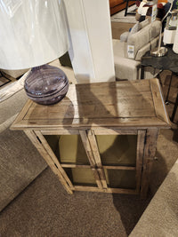 A Jofran Rustic Shores 2 Door Accent Cabinet with glass doors sits between two sofas, topped with a purple lamp.