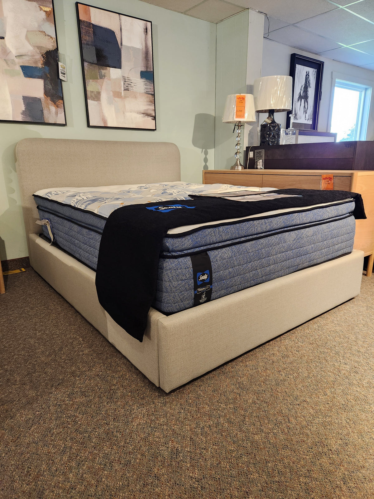 The Palliser EQ3 Nara Queen Bedframe with blue mattress and a beige upholstered headboard in a showroom.