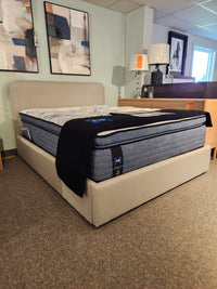 The Palliser EQ3 Nara Queen Bedframe with blue mattress and a beige upholstered headboard in a showroom.