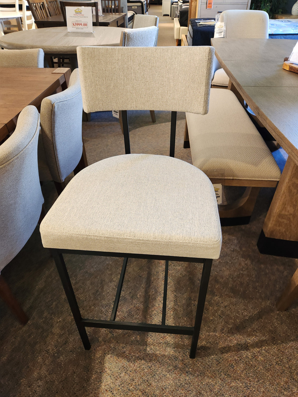 The Amisco Lucas Non Swivel Bar Stool with a metal frame and beige upholstery is displayed in the showroom.