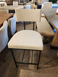 The Amisco Lucas Non Swivel Bar Stool with a metal frame and beige upholstery is displayed in the showroom.