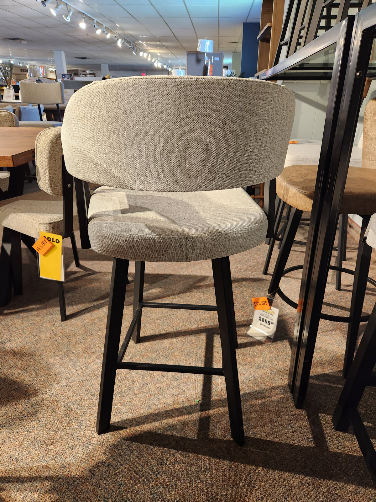 The Amisco Carmen Swivel Bar Stool in light gray with black legs is showcased in the showroom, price tags attached.