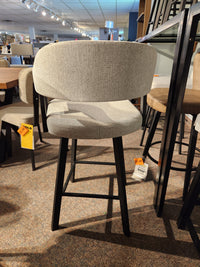 The Amisco Carmen Swivel Bar Stool in light gray with black legs is showcased in the showroom, price tags attached.