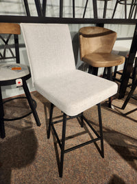 The Amisco Dorian Swivel Bar Stool features light gray upholstery and black metal legs, showcased in a showroom.