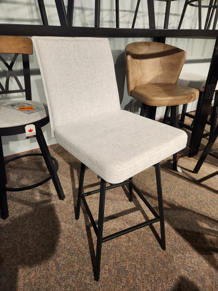 The Amisco Dorian Swivel Bar Stool features light gray upholstery and black metal legs, showcased in a showroom.