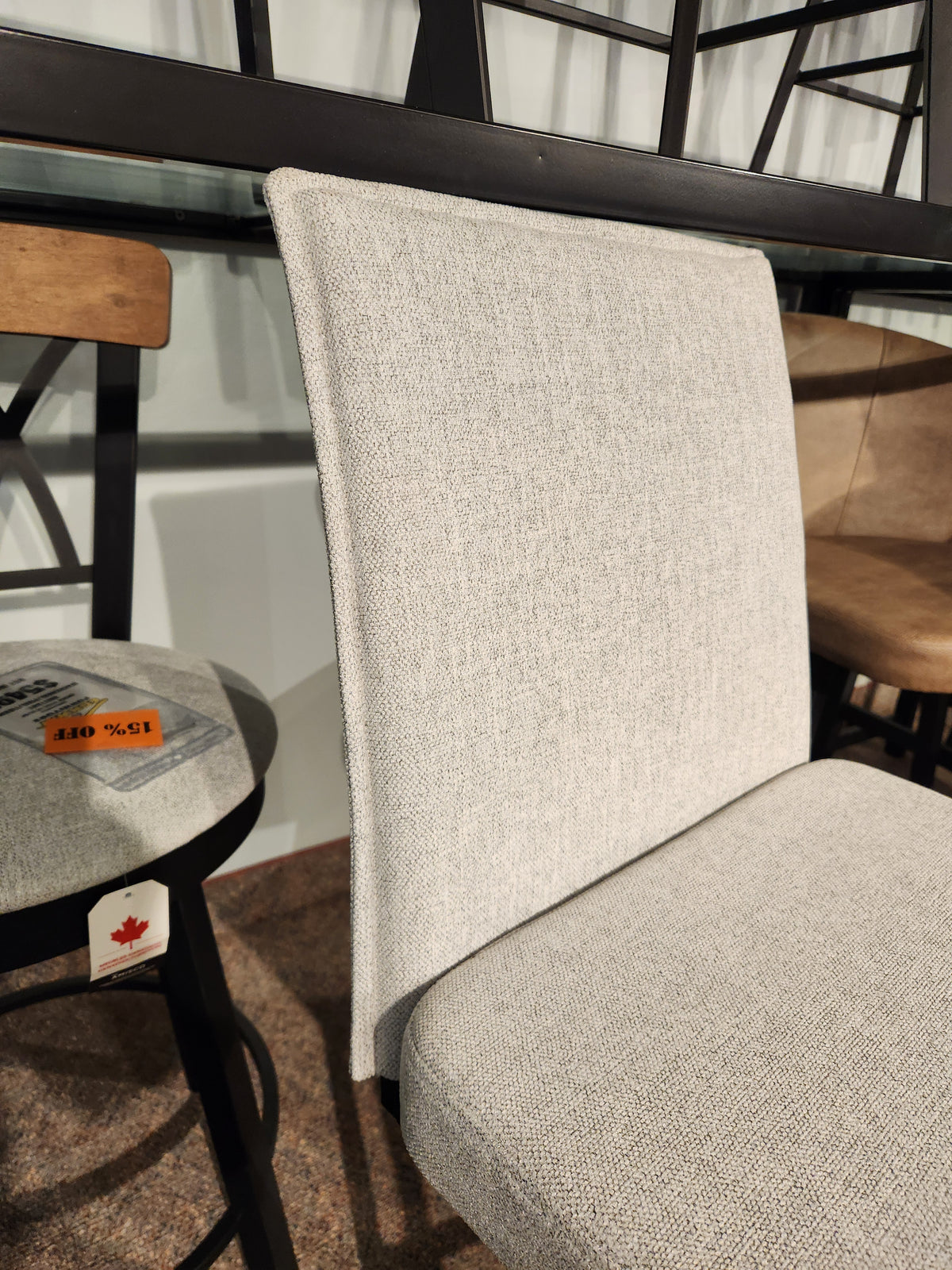 A light gray upholstered dining chair next to the Amisco Dorian Swivel Bar Stool with a sale tag in a showroom.