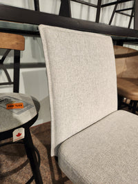 A light gray upholstered dining chair next to the Amisco Dorian Swivel Bar Stool with a sale tag in a showroom.