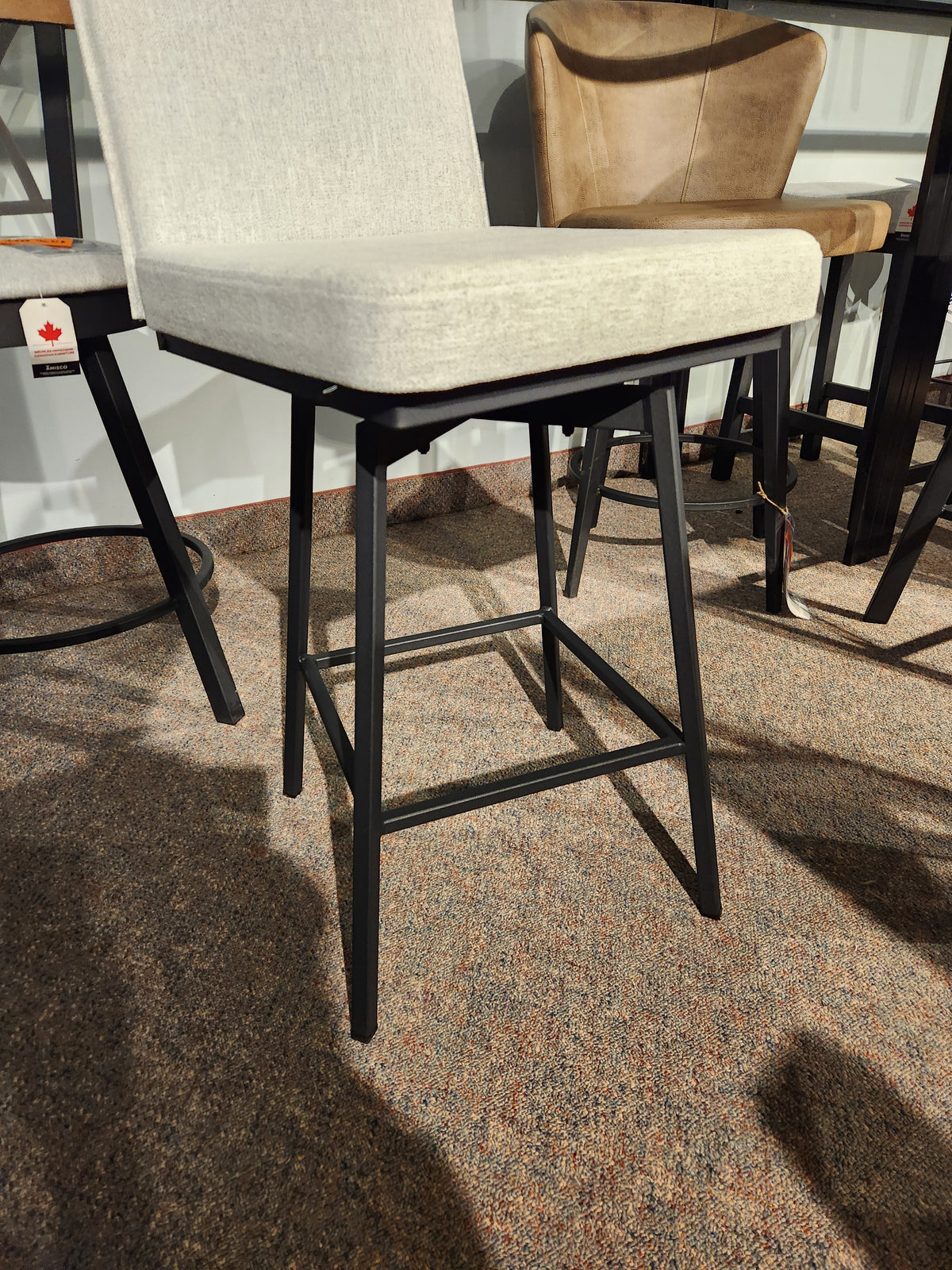 The Dorian Swivel Bar Stool by Amisco features a beige cushion and black legs, shown on a brown carpet among other chairs.