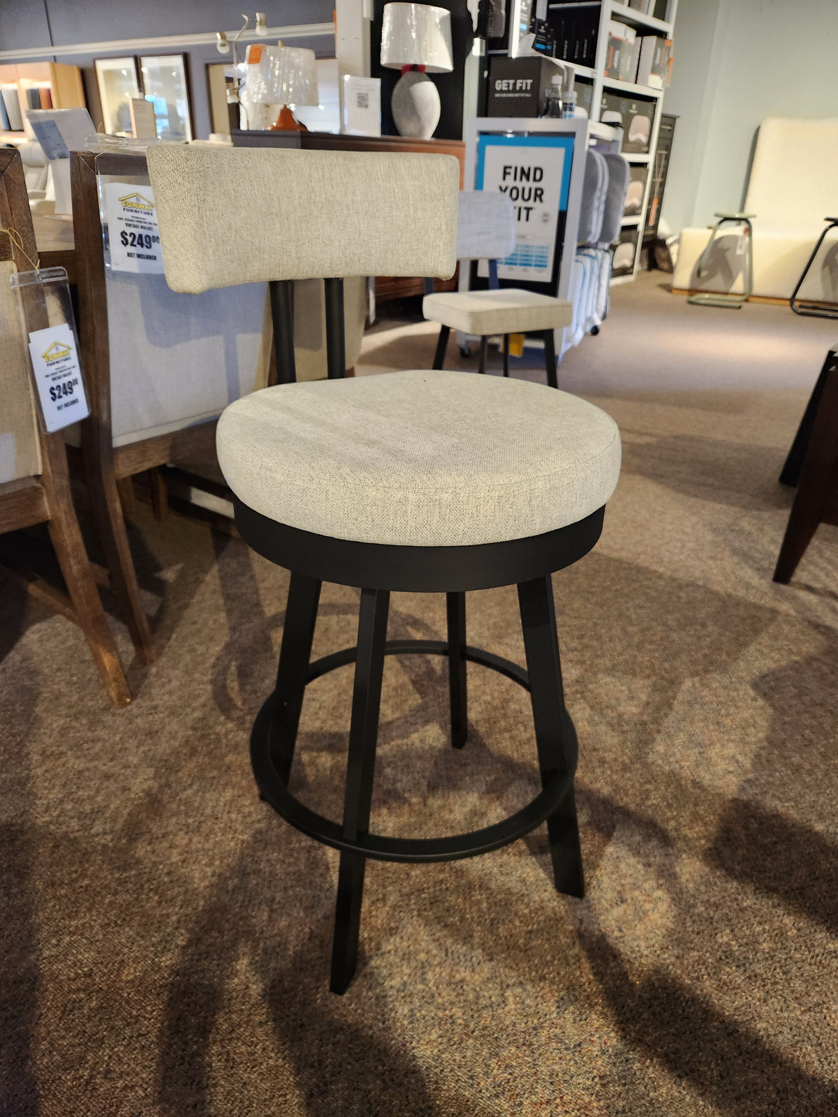 The Amisco Barry Swivel Bar Stool, beige with a black scratch-resistant finish, is on display in a furniture store.