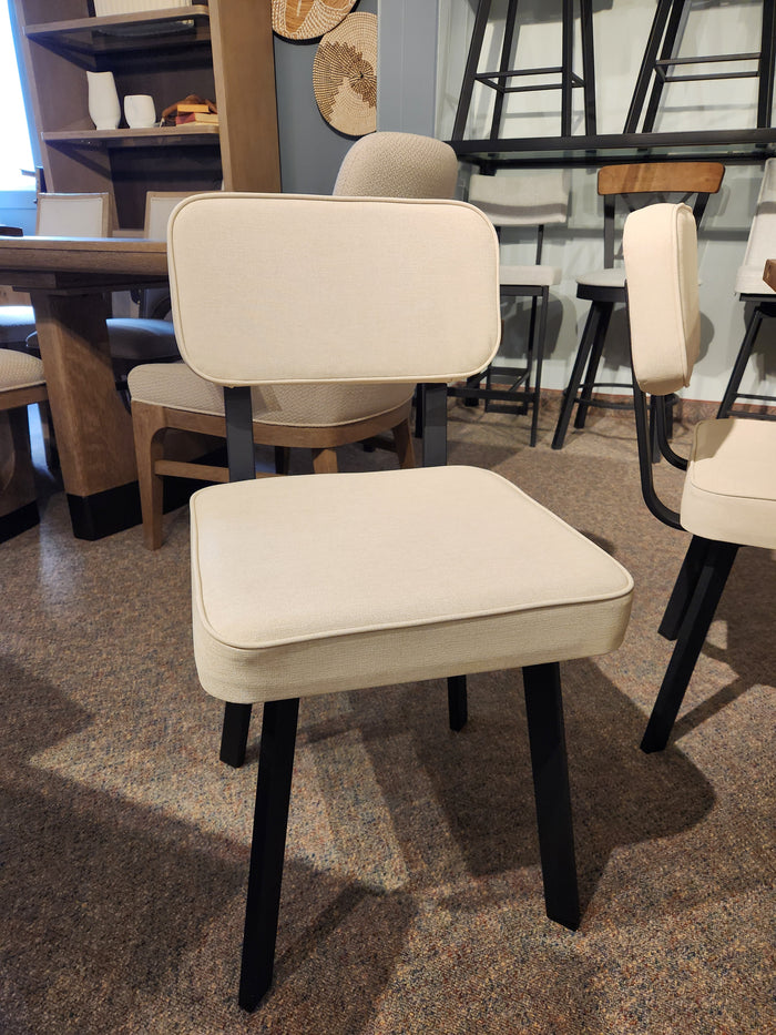 The Amisco Brixton Dining Chair, beige cushioned with black legs, is shown in a showroom among other chairs and tables.