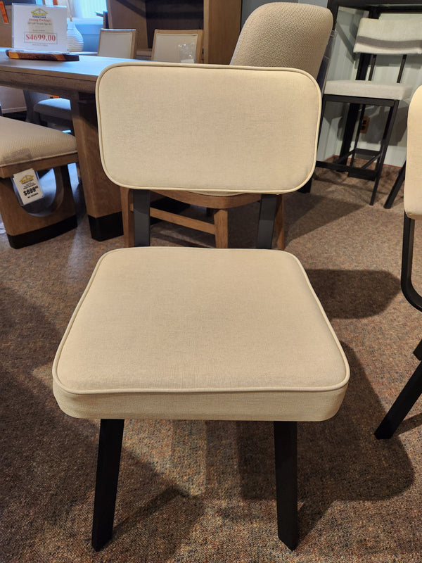 The Amisco Brixton Dining Chair, in beige upholstery with black legs, is displayed on a brown carpet in the store.