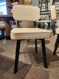 The Amisco Brixton Dining Chair, beige upholstered with a rectangular back and black legs, is shown in a showroom.