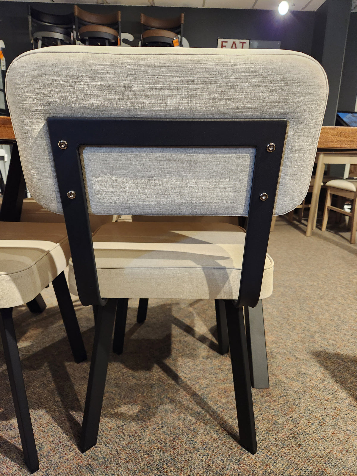 The Amisco Brixton Dining Chair in beige upholstery with black metal frame, shown from the back beside a wooden table.