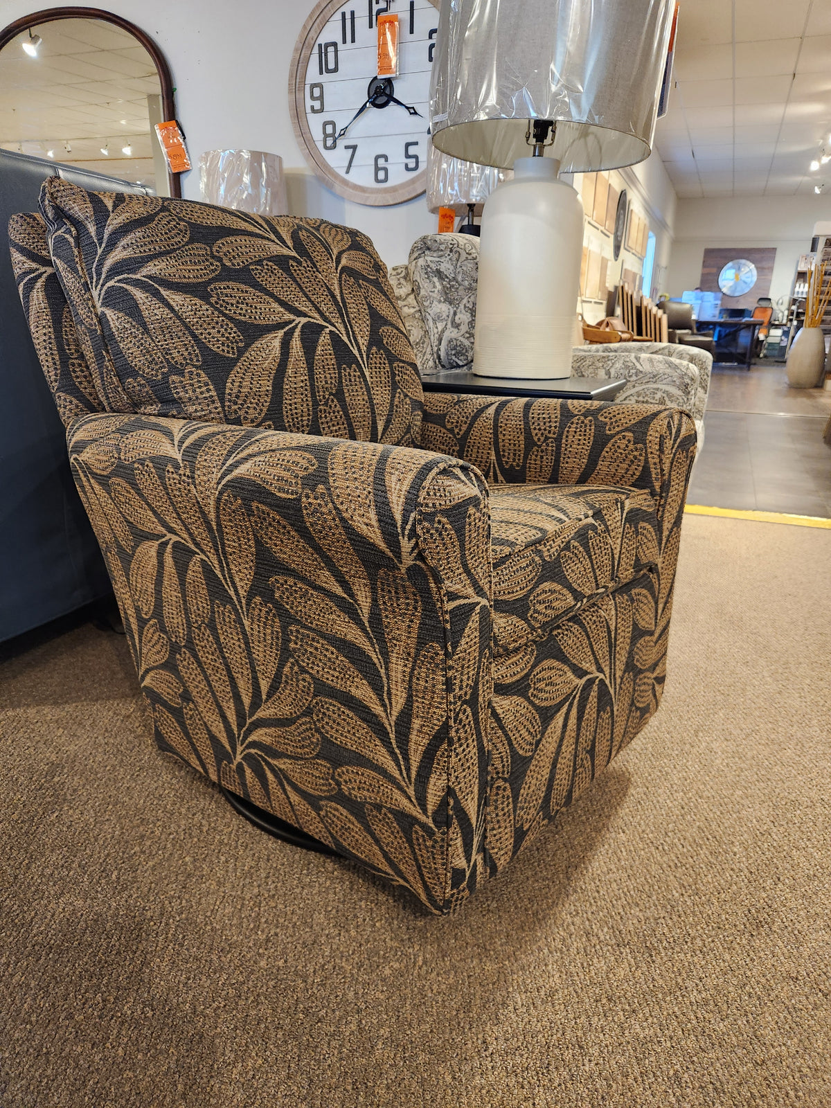 The Best Kacey Swivel Glider Chair with reversible cushion and dark leaf pattern is displayed on brown carpet in the showroom.
