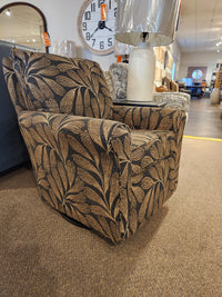 The Best Kacey Swivel Glider Chair with reversible cushion and dark leaf pattern is displayed on brown carpet in the showroom.