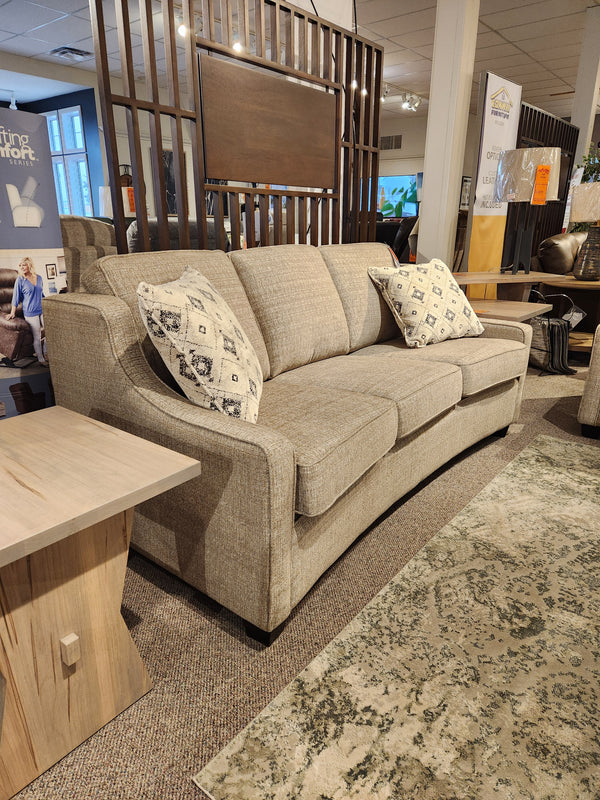 The Superstyle 9670 Sofa in beige, medium-firm, with two throw pillows is shown beside a wooden side table in a showroom.