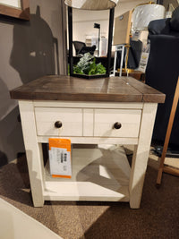 A Jofran 1706 Madison County End Table with barn door accents and a drawer is displayed in a furniture store.