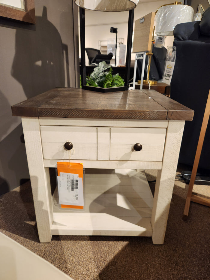 A Jofran 1706 Madison County End Table with barn door accents and a drawer is displayed in a furniture store.
