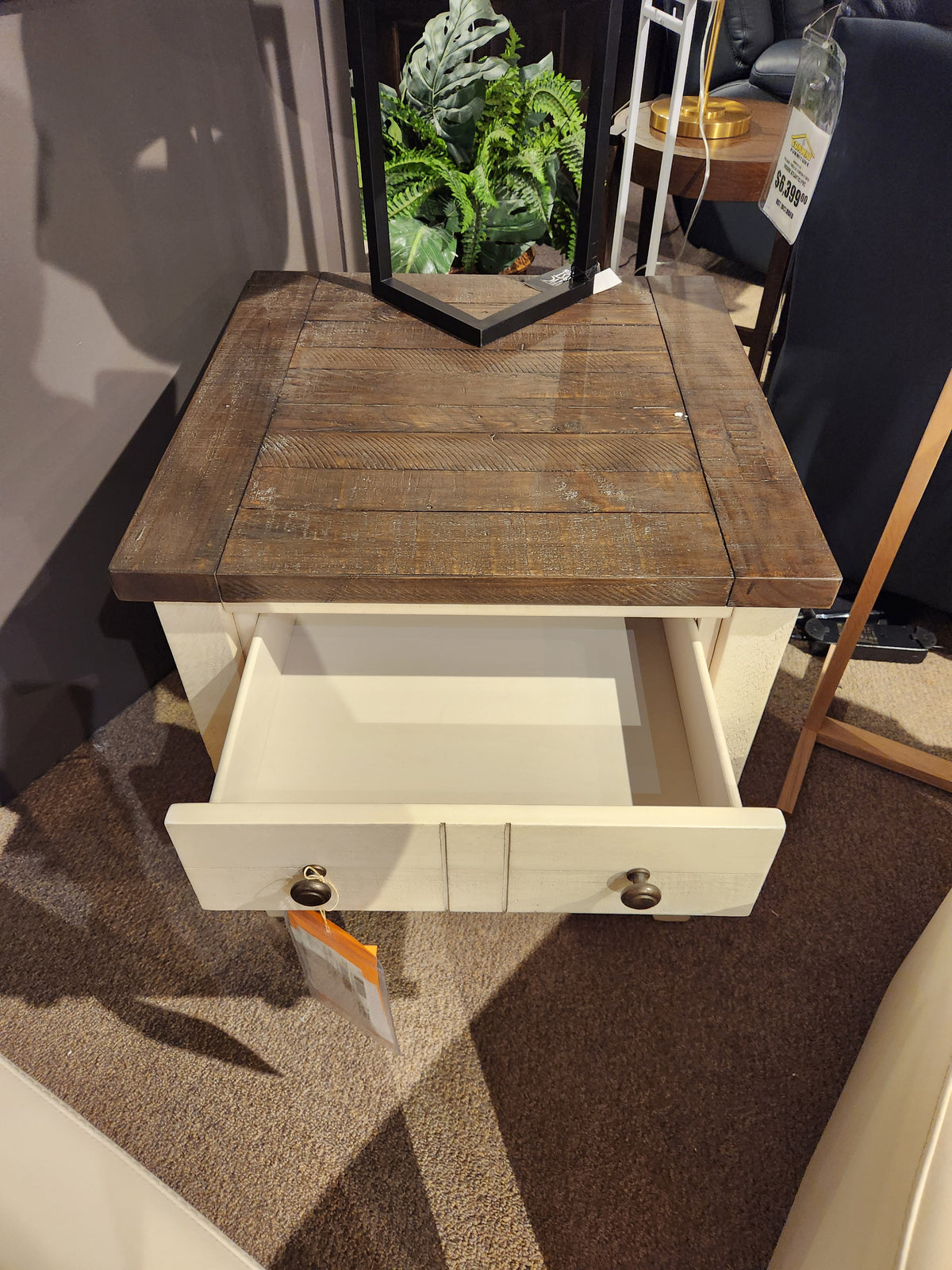 The Jofran 1706 Madison County End Table features barn door accents, a white drawer, and plant decor on top.