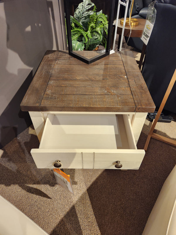 The Jofran 1706 Madison County End Table features barn door accents, a white drawer, and plant decor on top.