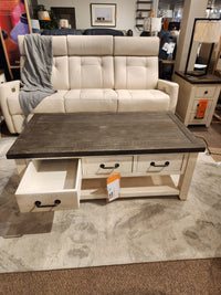 Beige sofa behind a Jofran 1706 Madison County Coffee Table with barn door accents in a furniture store.