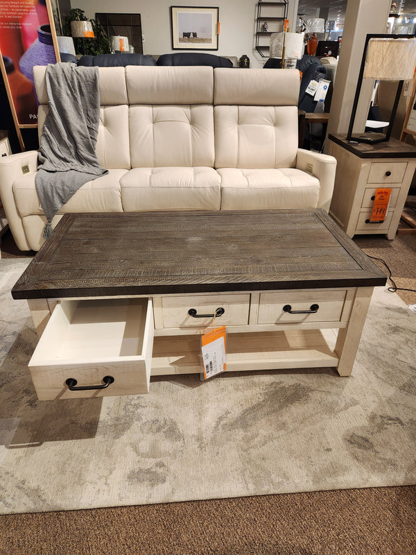Beige sofa behind a Jofran 1706 Madison County Coffee Table with barn door accents in a furniture store.